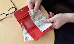 Ręce wkładają banknot 500 zł do czerwonego portfela. Obok leżą okulary.