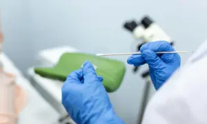 Ręce w niebieskich rękawiczkach przygotowują próbkę do badań cytologia wyniki. W tle mikroskop i fotel ginekologiczny.