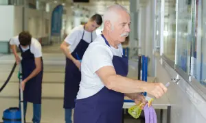 Starszy pan z wąsami i dwaj młodsi mężczyźni sprzątają korytarz. Zastanawiają się, ile można dorobić do emerytury, pracując w ten sposób.