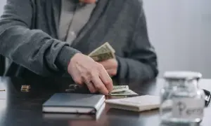 Starszy mężczyzna liczy pieniądze, zastanawiając się, czy renta wlicza się do emerytury. Obok leży skarbonka.