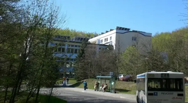 Sanatorium uzdrowiskowe „Leśnik” otoczone zielenią, z autobusem na pierwszym planie i ludźmi czekającymi na przystanku.