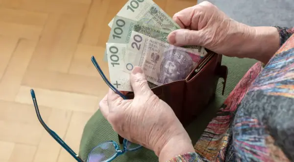 Dłoń starszej osoby wyjmuje z portfela polskie banknoty. Może to symbolizować dodatek do emerytury po 75 roku życia.