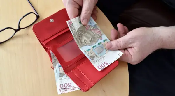 Ręce wkładają banknot 500 zł do czerwonego portfela. Obok leżą okulary.