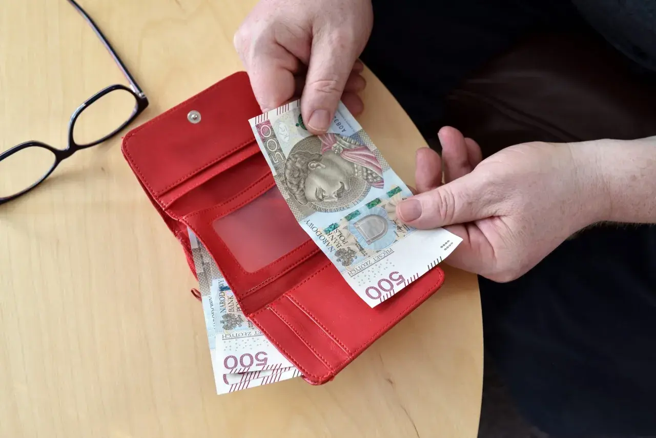 Ręce wkładają banknot 500 zł do czerwonego portfela. Obok leżą okulary.
