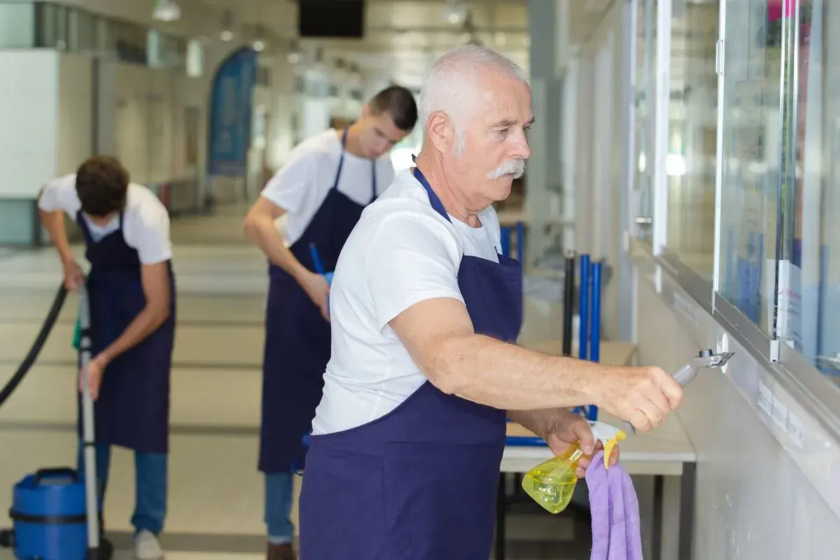 Starszy pan z wąsami i dwaj młodsi mężczyźni sprzątają korytarz. Zastanawiają się, ile można dorobić do emerytury, pracując w ten sposób.