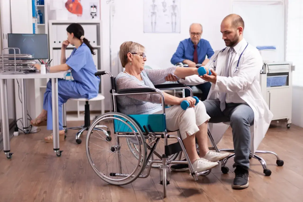 Rehabilitacja po udarze: starsza pani w wózku ćwiczy z fizjoterapeutą, który pomaga jej podnosić hantle. W tle pielęgniarka i pacjent.