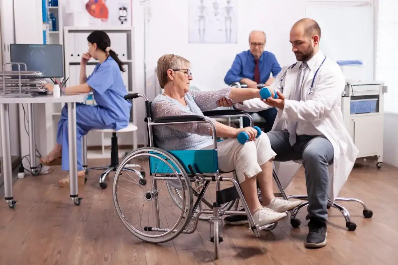 Rehabilitacja po udarze: starsza pani w wózku ćwiczy z fizjoterapeutą, który pomaga jej podnosić hantle. W tle pielęgniarka i pacjent.