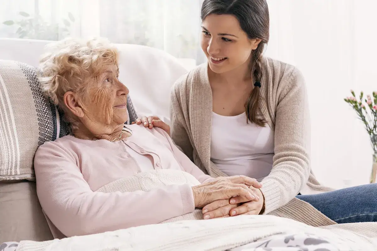 Młoda kobieta wspiera starszą panią, która zmaga się z chorobą Alzheimera. Ich dłonie splecione, pełne troski i zrozumienia.